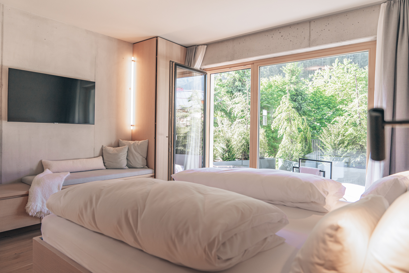 Doppelbett im Hotelzimmer mit großem Fernseher, gemütlicher Sitzecke und Balkon mit großer Glasfront